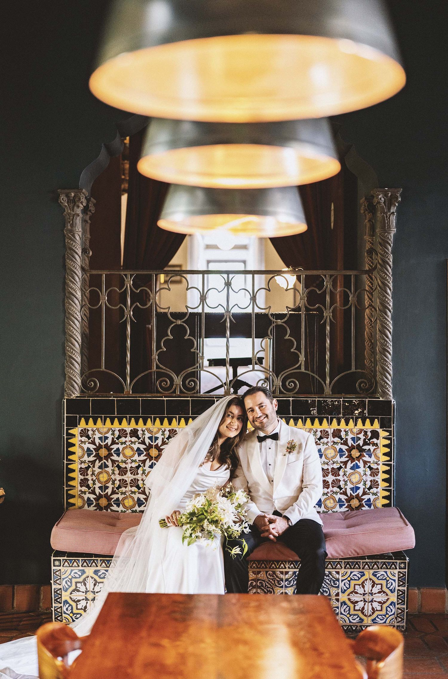 Bride and groom sitting together on a tiled bench under warm pendant lights in an elegant indoor wedding venue.