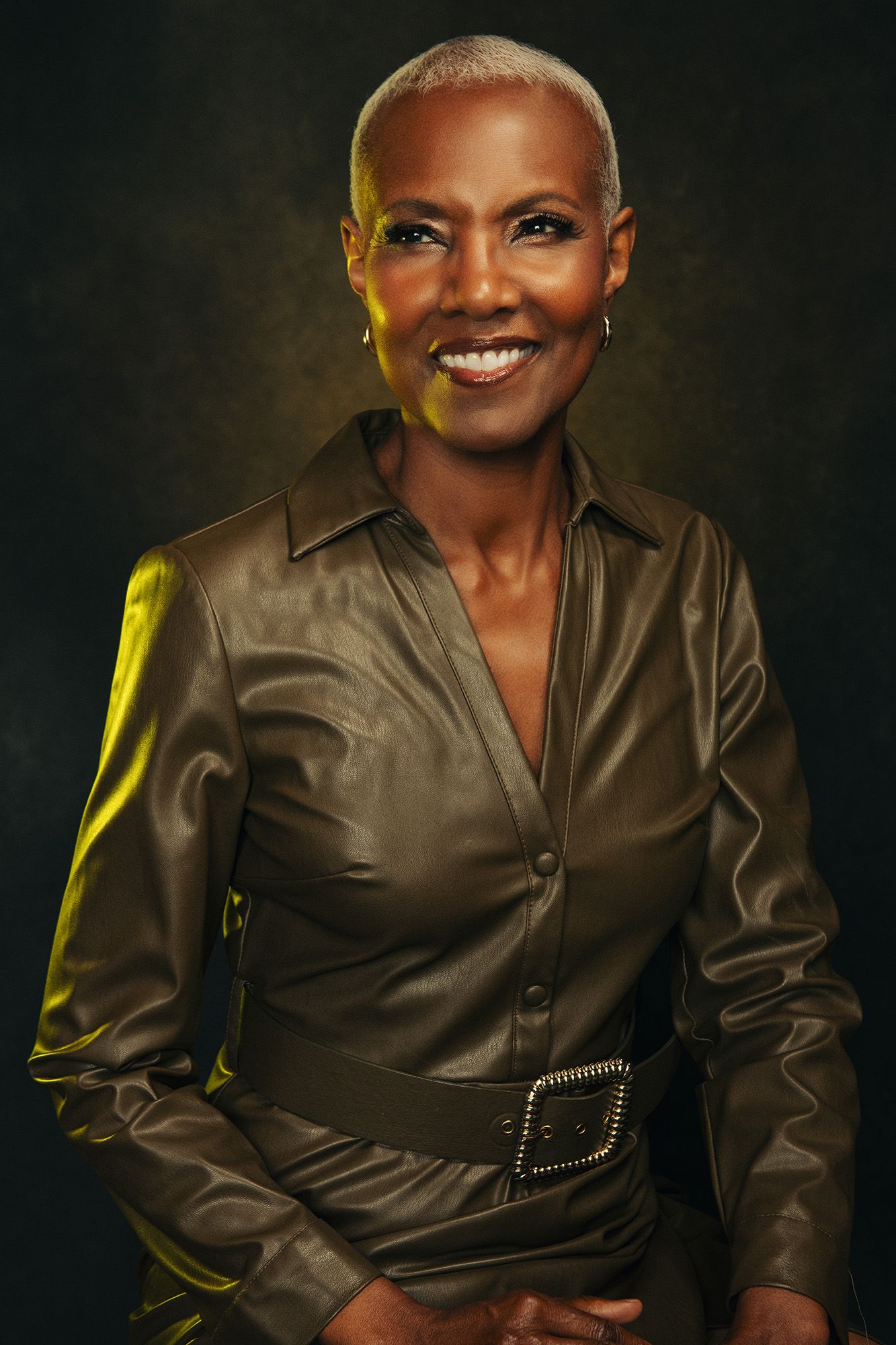 Smiling woman in olive leather belted shirt dress with short hair against dark background.