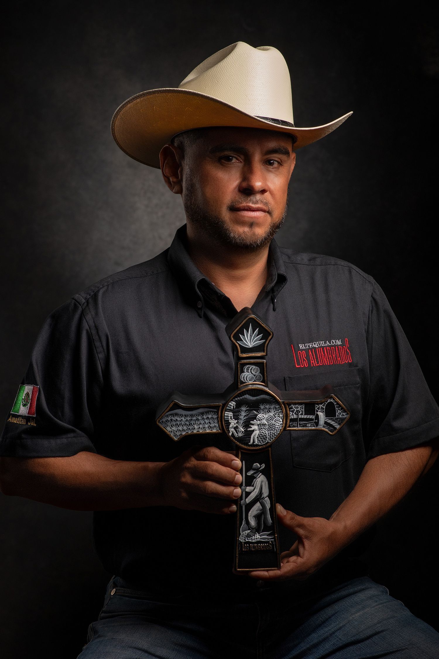 Man in black shirt and cowboy hat holds decorative cross trophy in dramatic studio portrait.