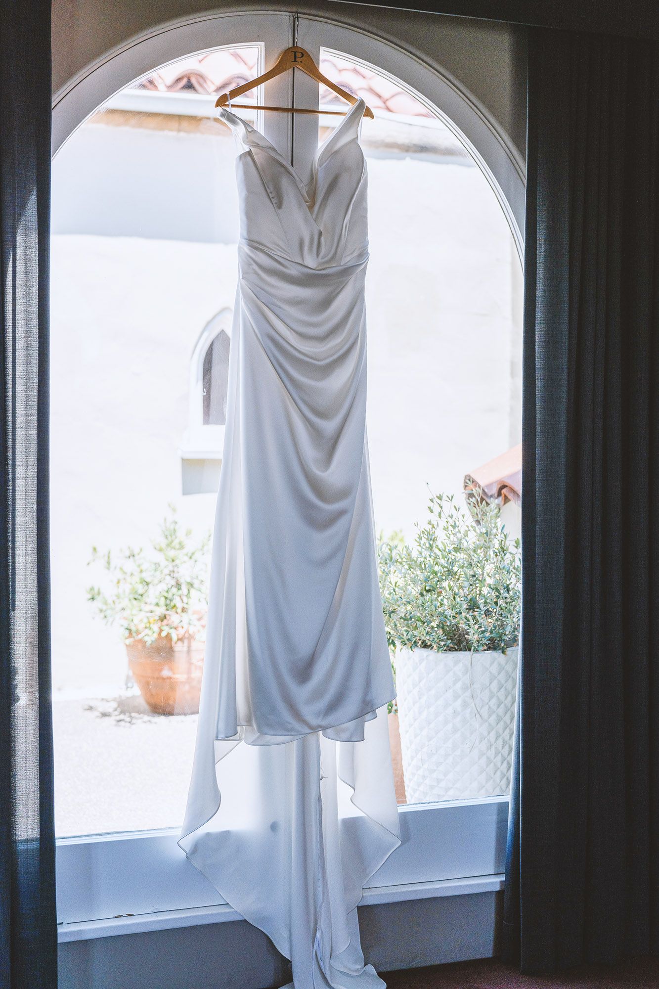 Elegant white wedding gown hanging on a wooden hanger in an arched window with natural light.