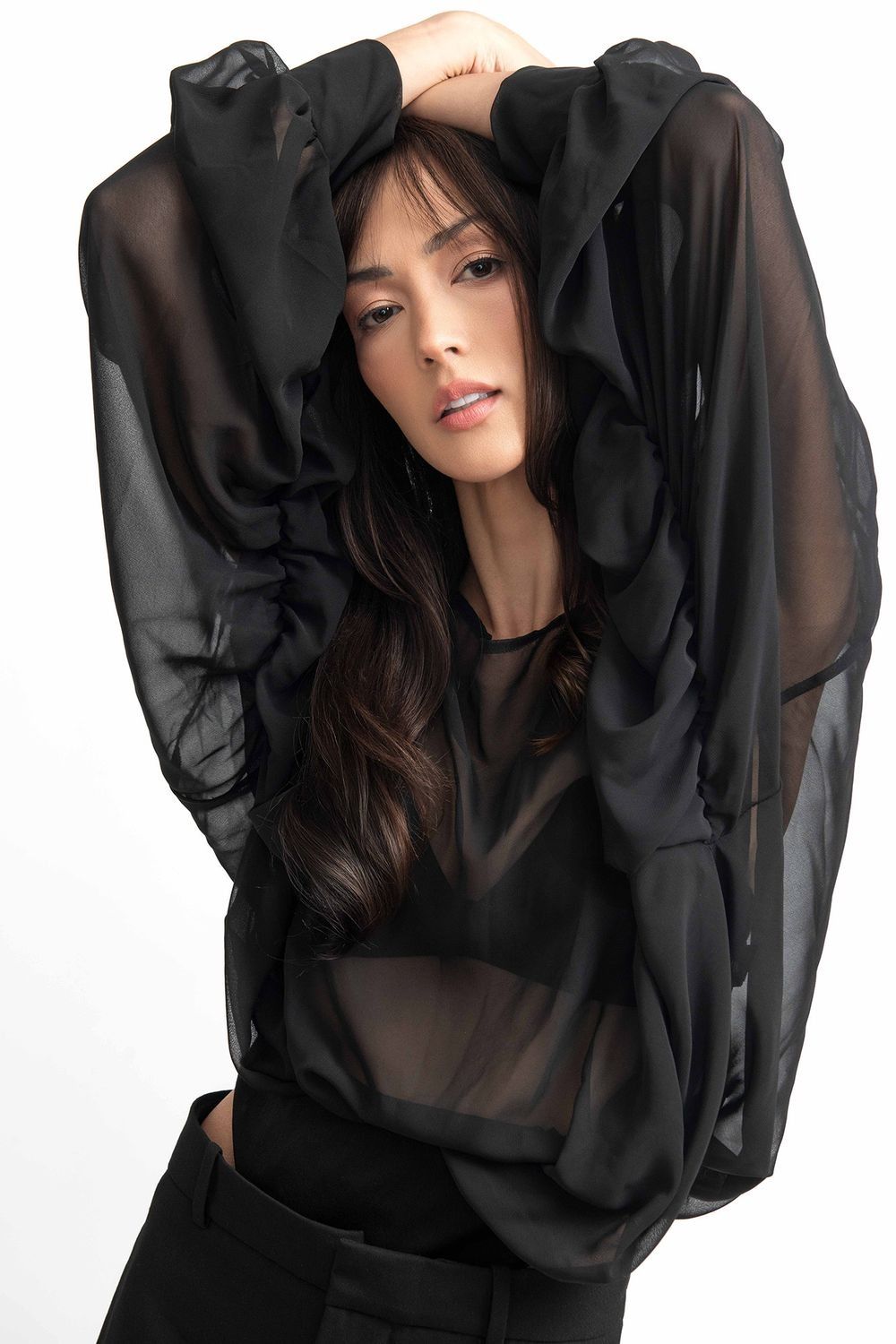 Brunette model posing in sheer black blouse with arms raised in minimalist studio shot.