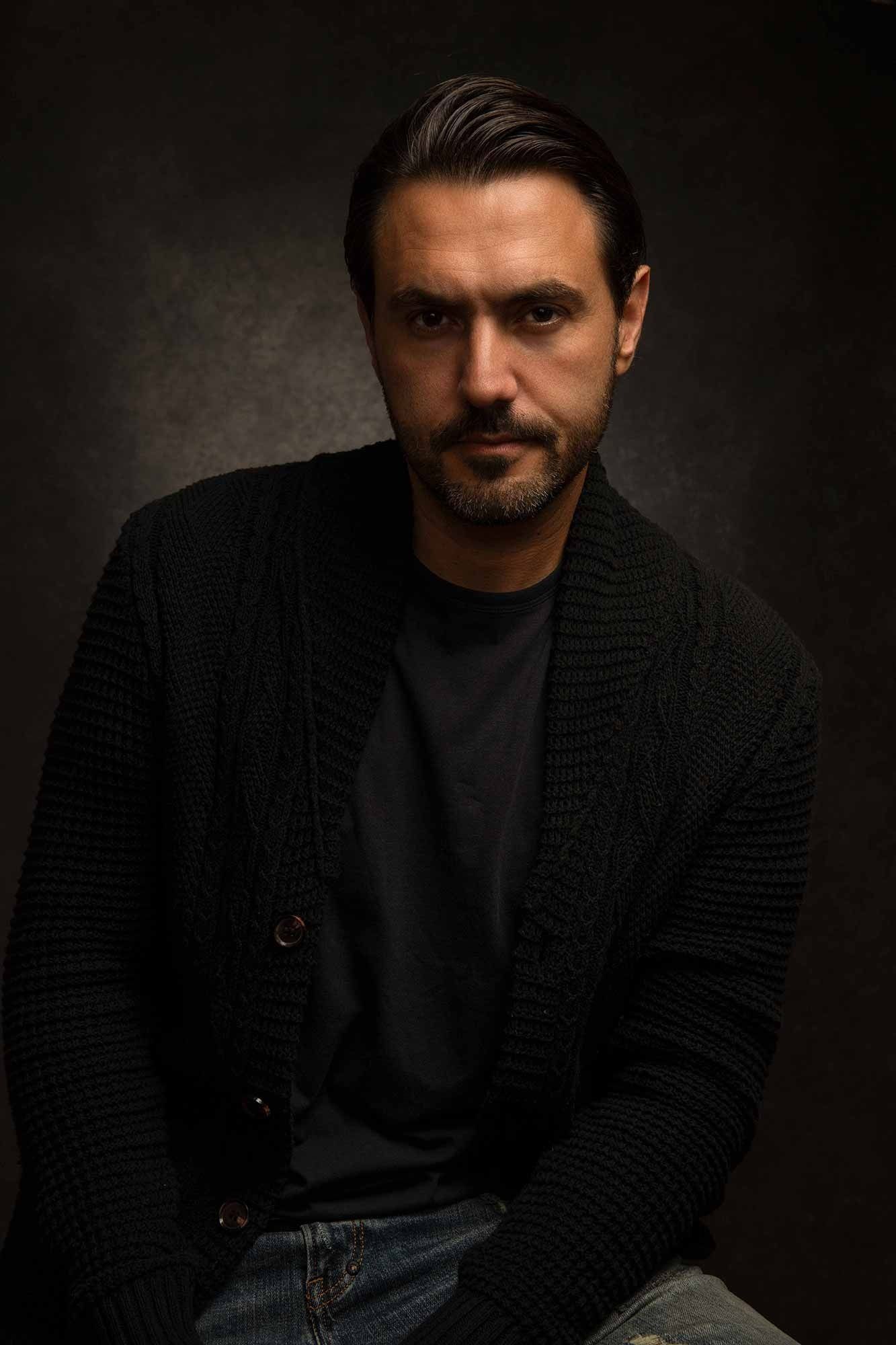 Bearded man in black knit cardigan seated in moody dark studio portrait photography.