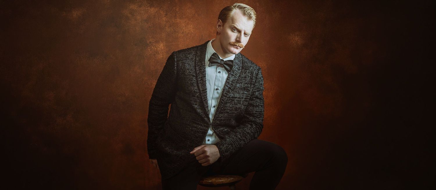 Elegant man in dark plaid suit with bow tie sitting against moody brown background.