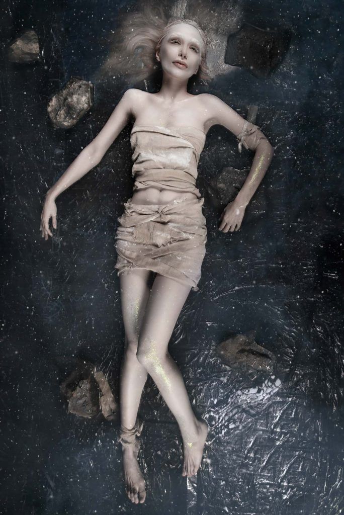 Artistic fashion photo of woman in beige draped mini dress lying against dark textured background.