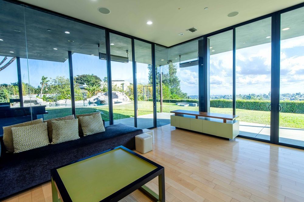 Modern living room with floor-to-ceiling glass walls, hardwood floors, blue sofa, and lush garden views.