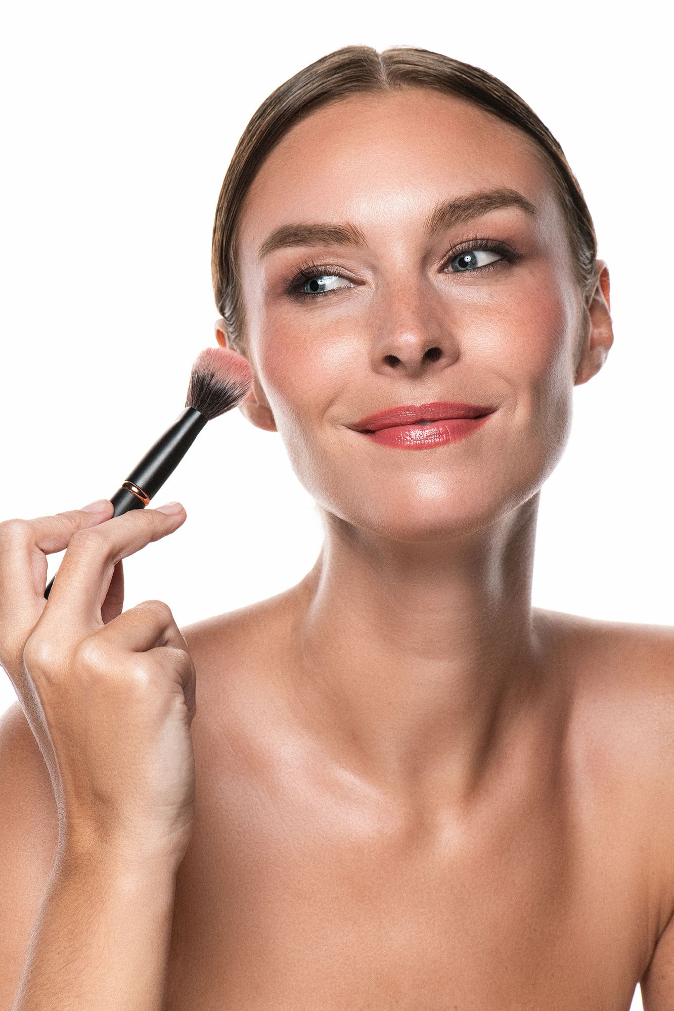 Smiling woman applying blush with makeup brush on white background, showcasing natural beauty look.