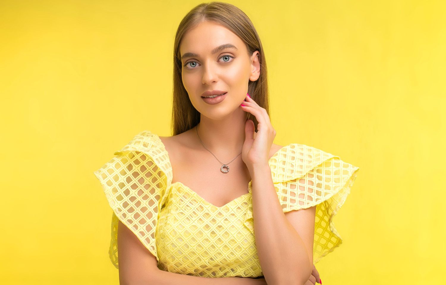 Blonde woman in yellow eyelet ruffle-shoulder top touching her ear against bright yellow background.