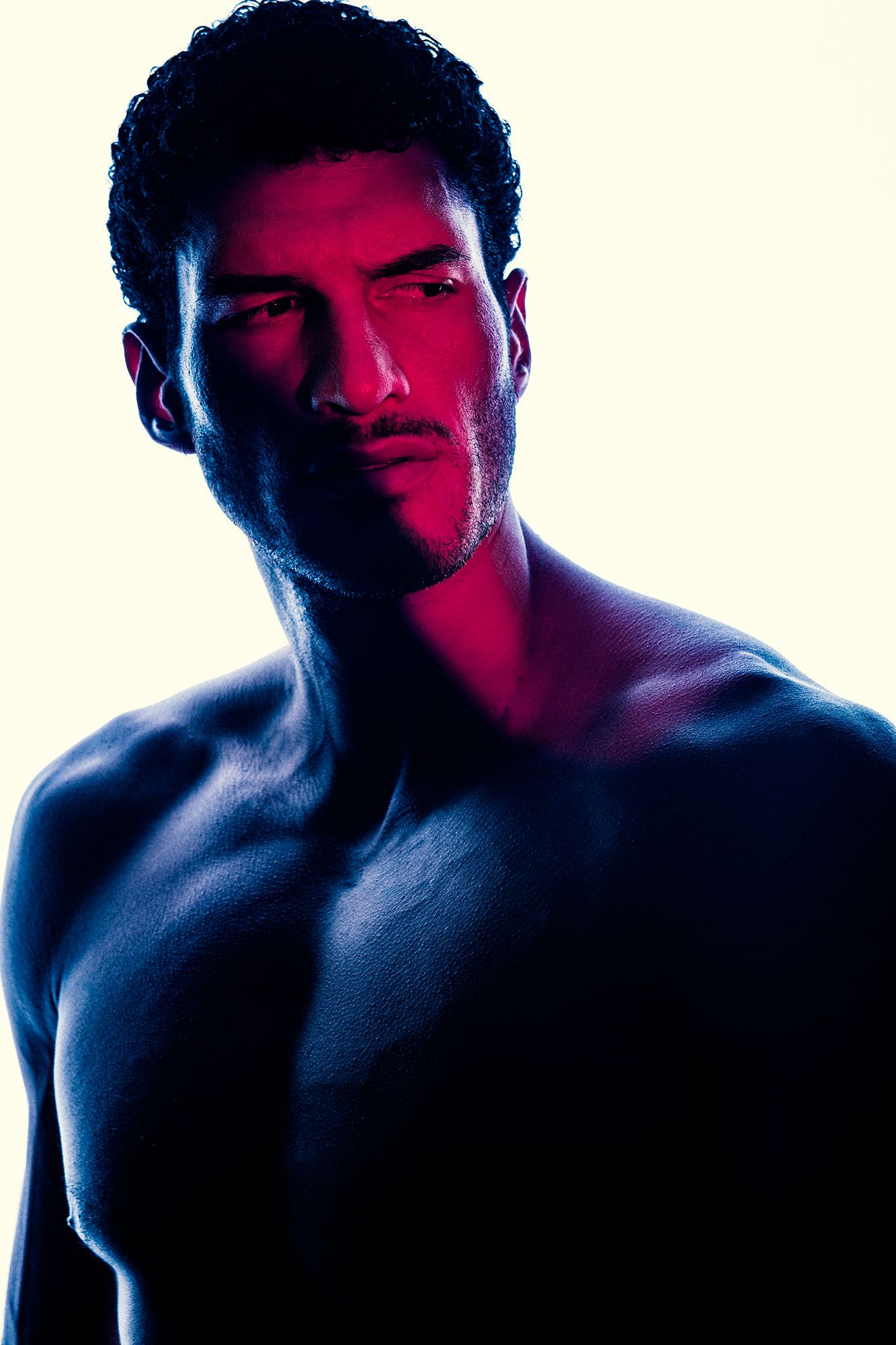 Dramatic portrait of a shirtless male model lit with blue and red lighting on white background.