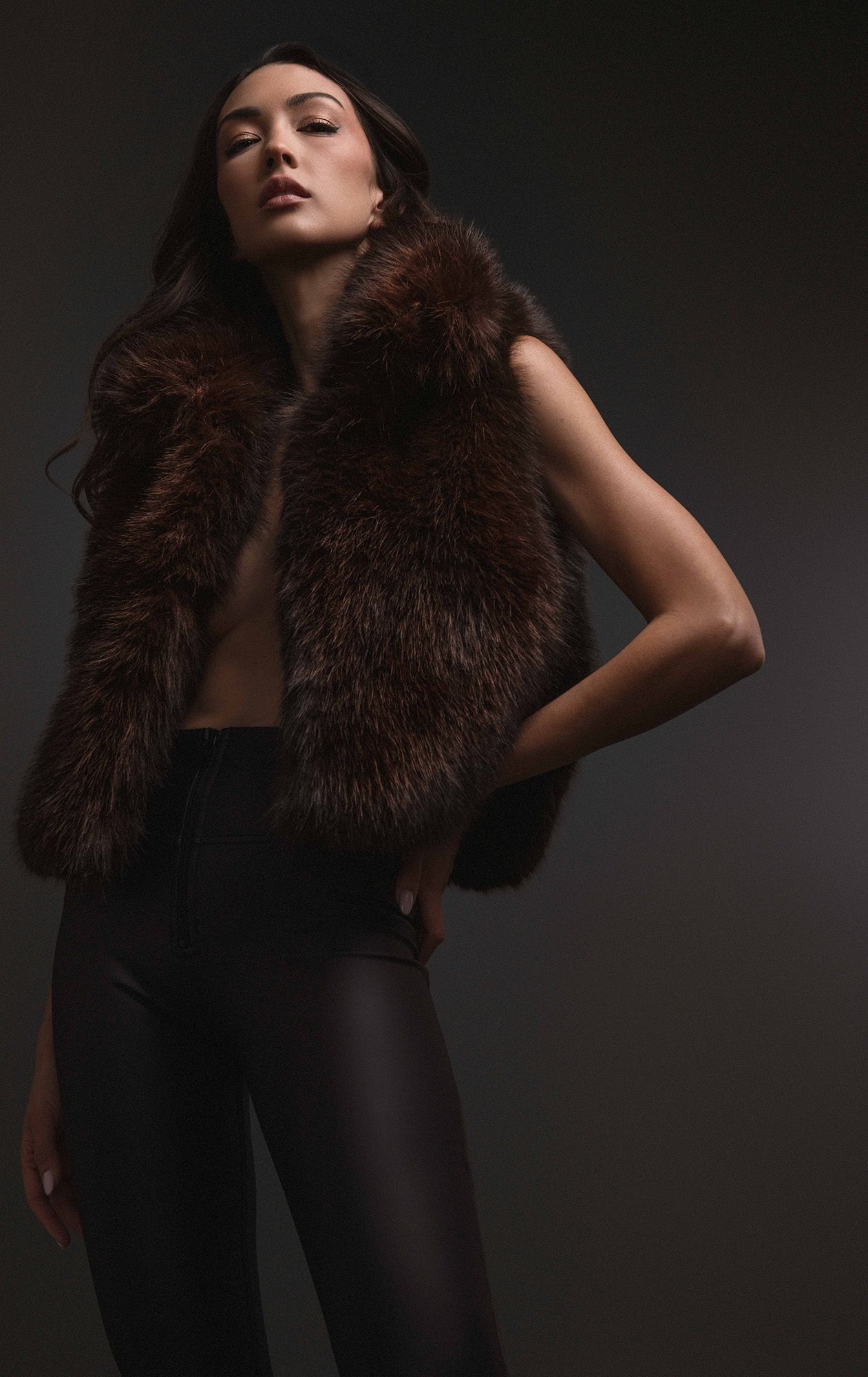 A tall brunette model wearing a brown fur vest and black leather pants in a fashion studio shoot.