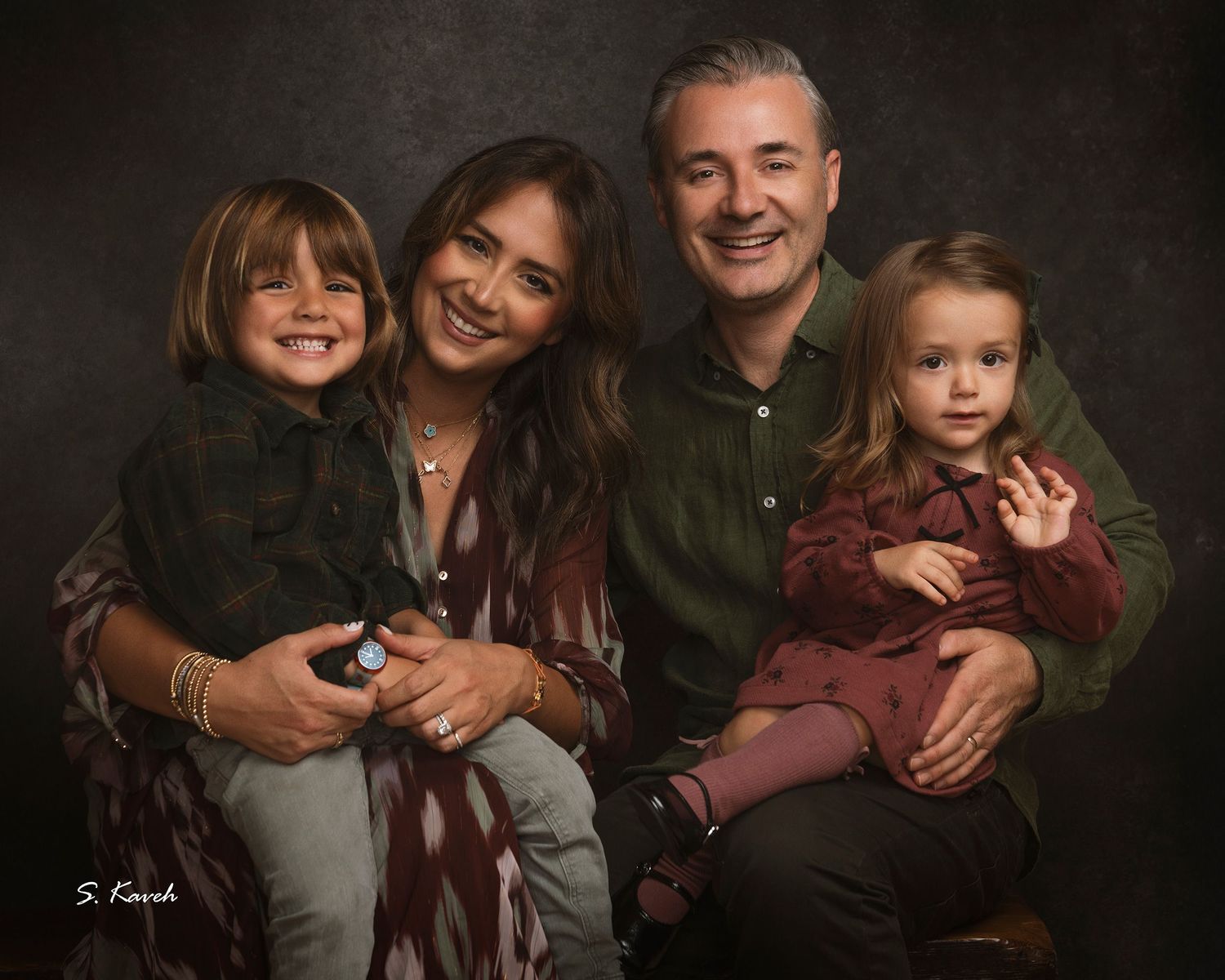 Smiling family portrait of two parents holding a young boy and a toddler girl against a dark textured studio backdrop.