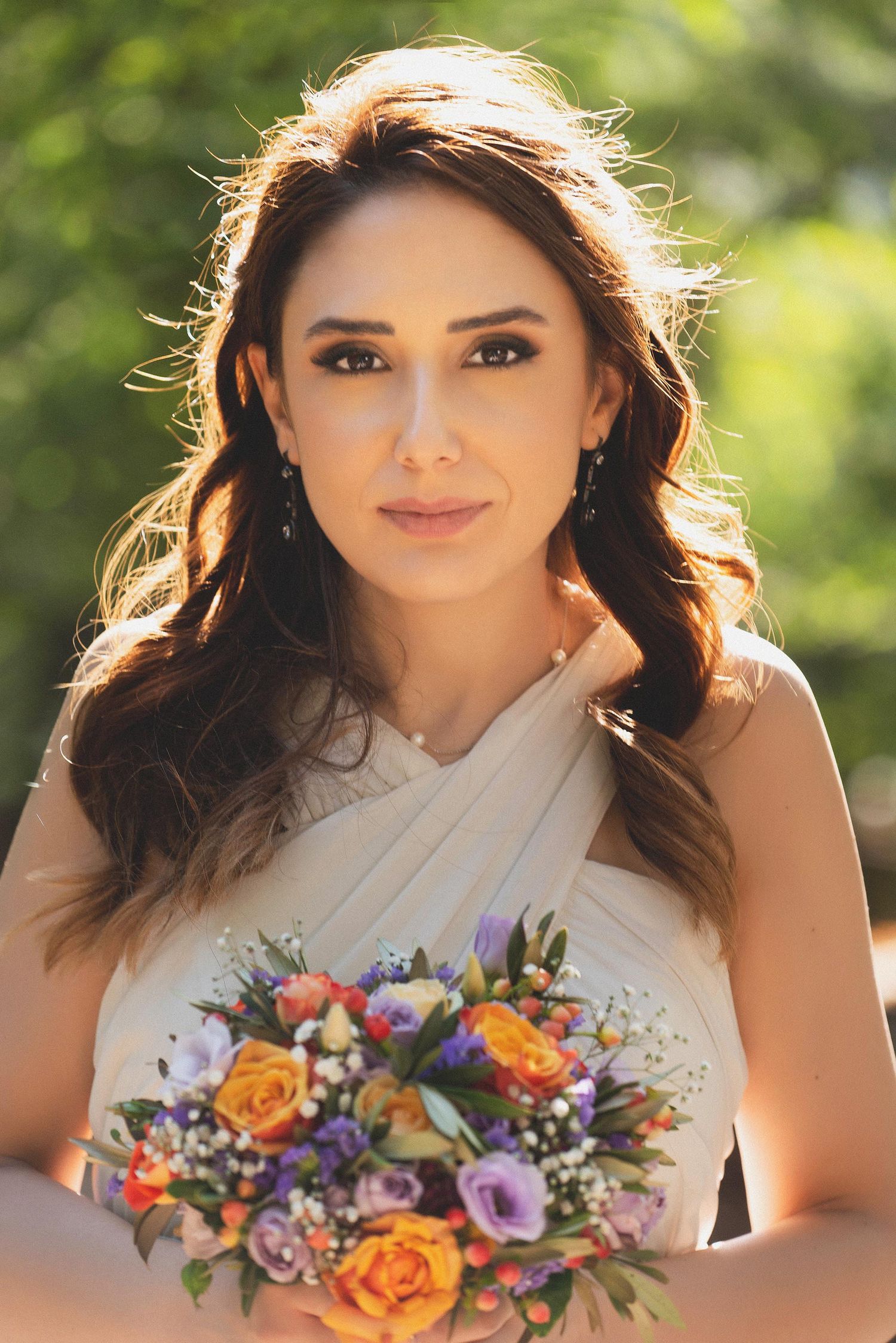 A beautiful bride in a white dress holds a colorful bouquet of roses and wildflowers outdoors in soft sunlight.