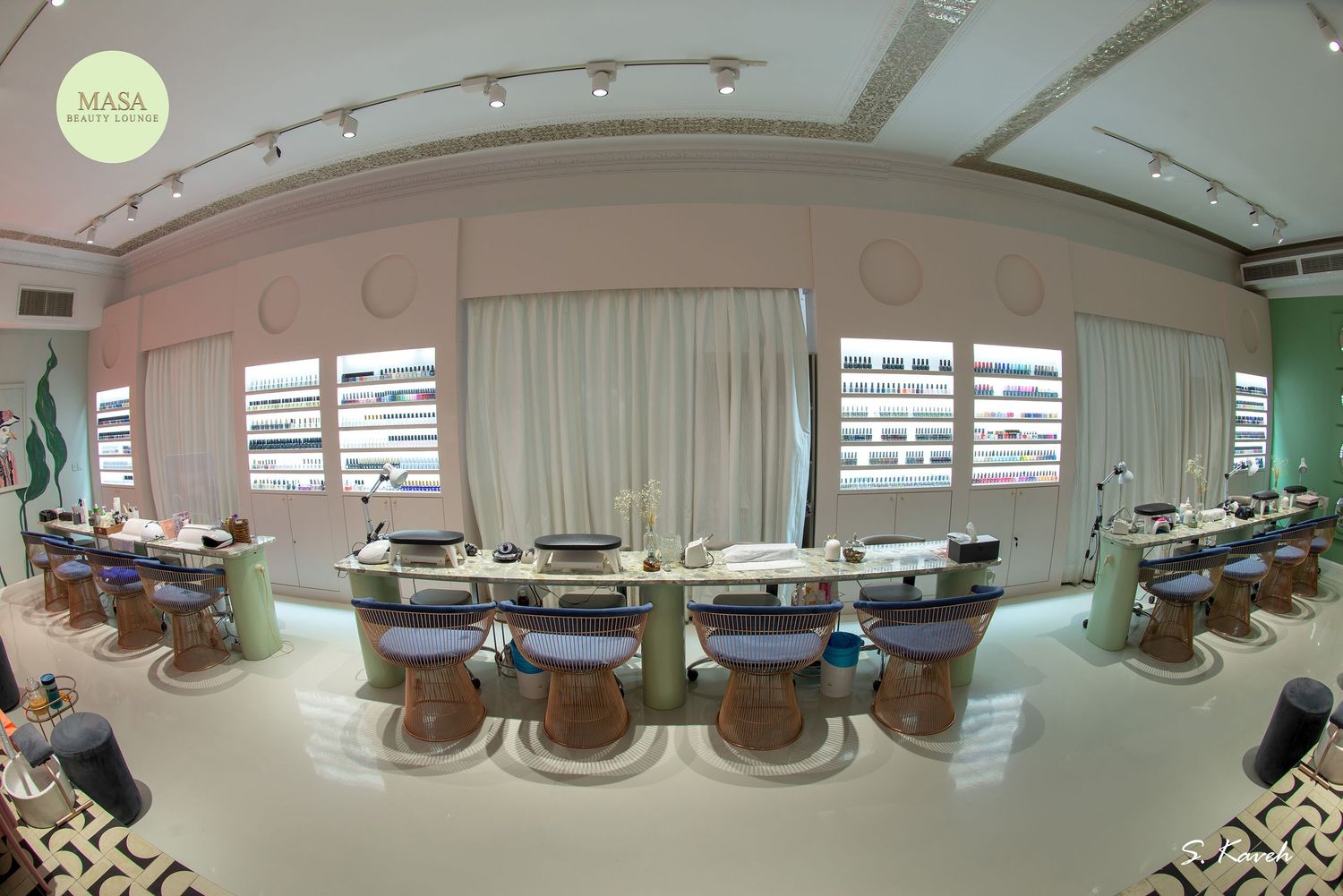 Elegant nail salon interior with long manicure tables, pink walls, white curtains, and modern lighting in a wide-angle view.