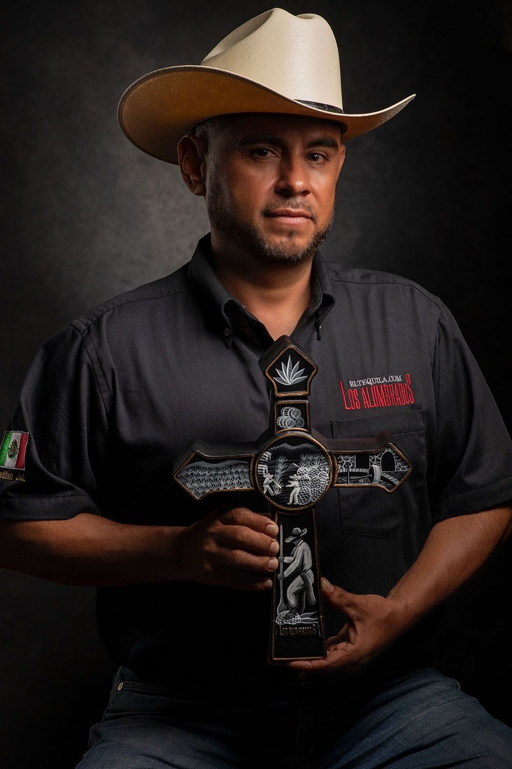 A Hispanic man in a cowboy hat holding a decorative cross wearing a black embroidered shirt in a studio portrait.