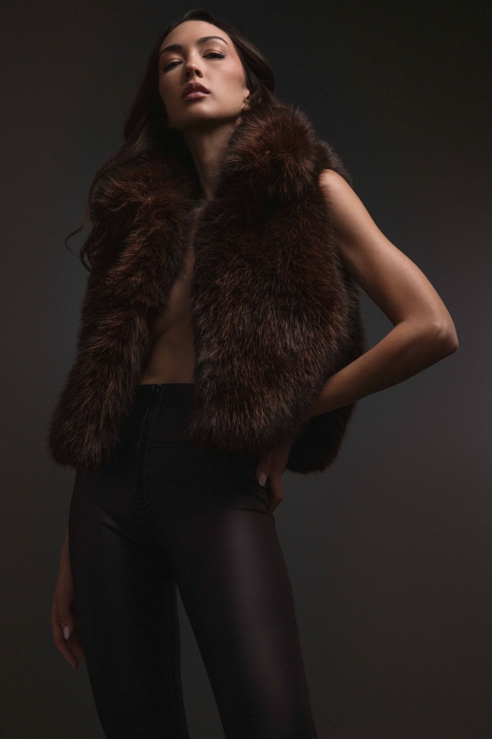 Woman wearing brown faux fur vest with black leather pants on dark background.