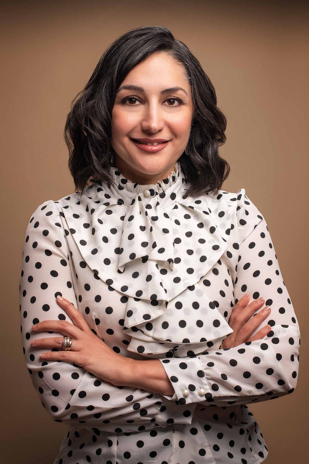 Smiling woman in polka dot blouse with arms crossed posing confidently against a warm brown background.
