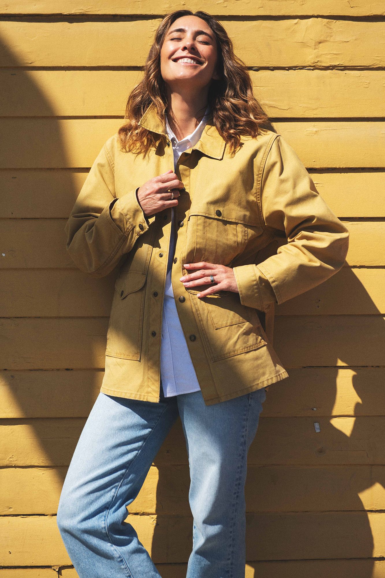 Woman smiling in yellow jacket and blue jeans leaning against a yellow wall in sunlight.