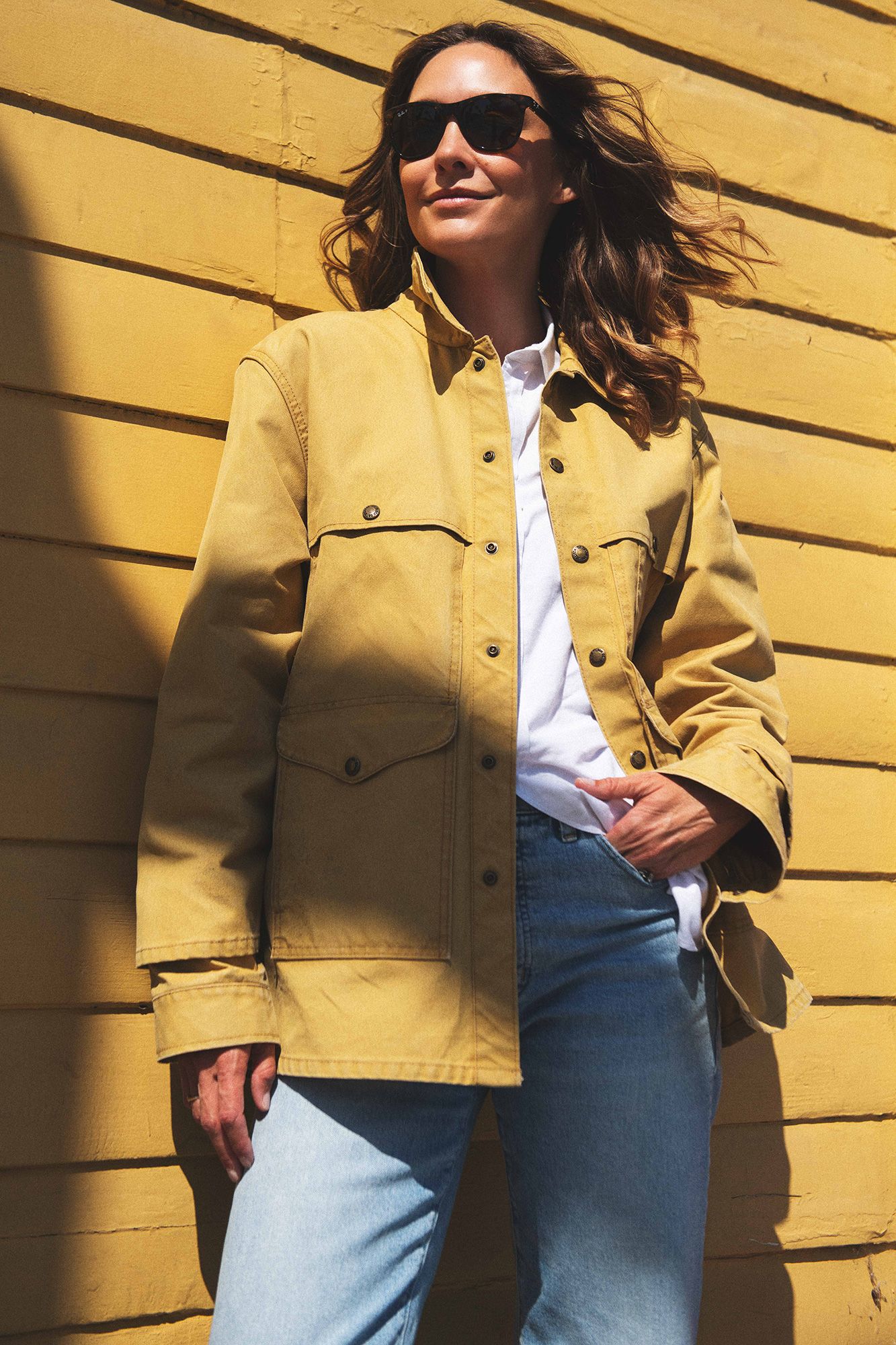 Woman in yellow jacket, white shirt, and jeans poses against a yellow wall wearing sunglasses in sunlight.