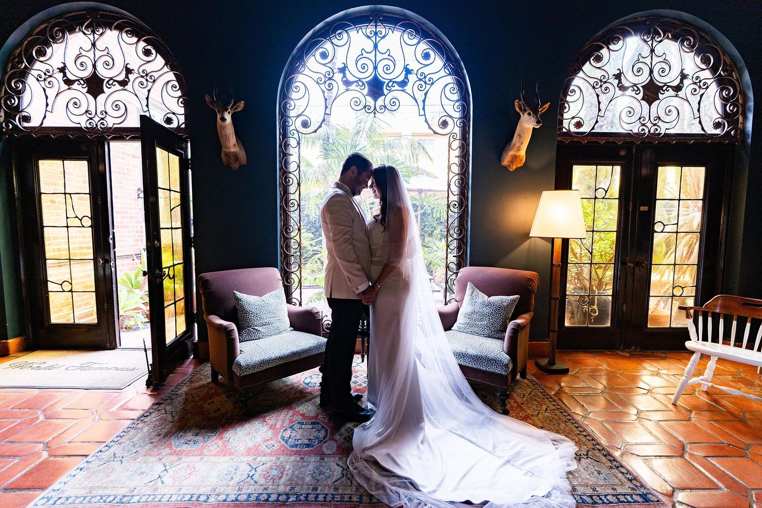 Bride and groom share romantic kiss in elegant venue with ornate arched windows and stained glass.