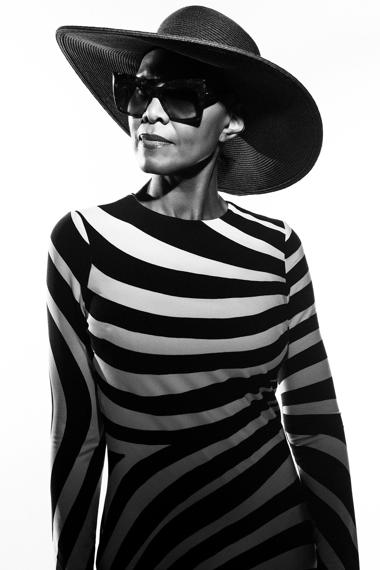 Stylish woman in striped dress with wide-brim black hat and sunglasses in black and white photo.