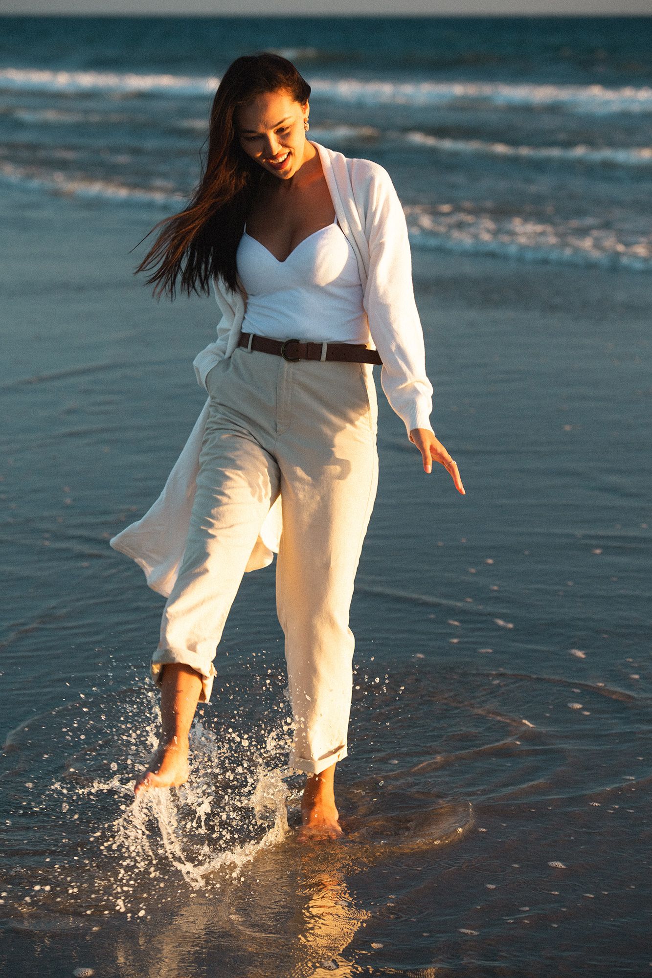 Woman in white outfit splashing barefoot in ocean waves at sunset on a sandy beach.