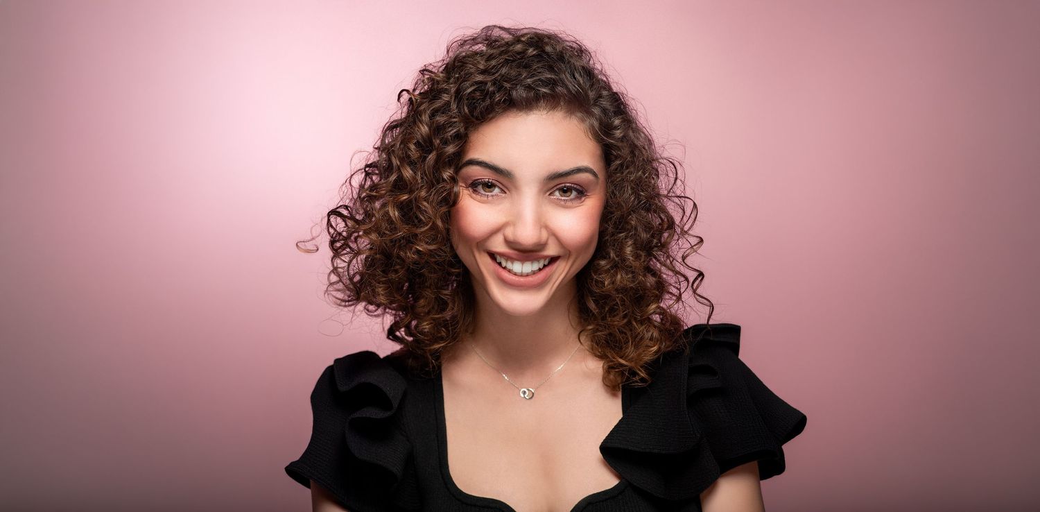 Smiling young woman with curly brown hair wearing a black ruffled top against a soft pink studio background.