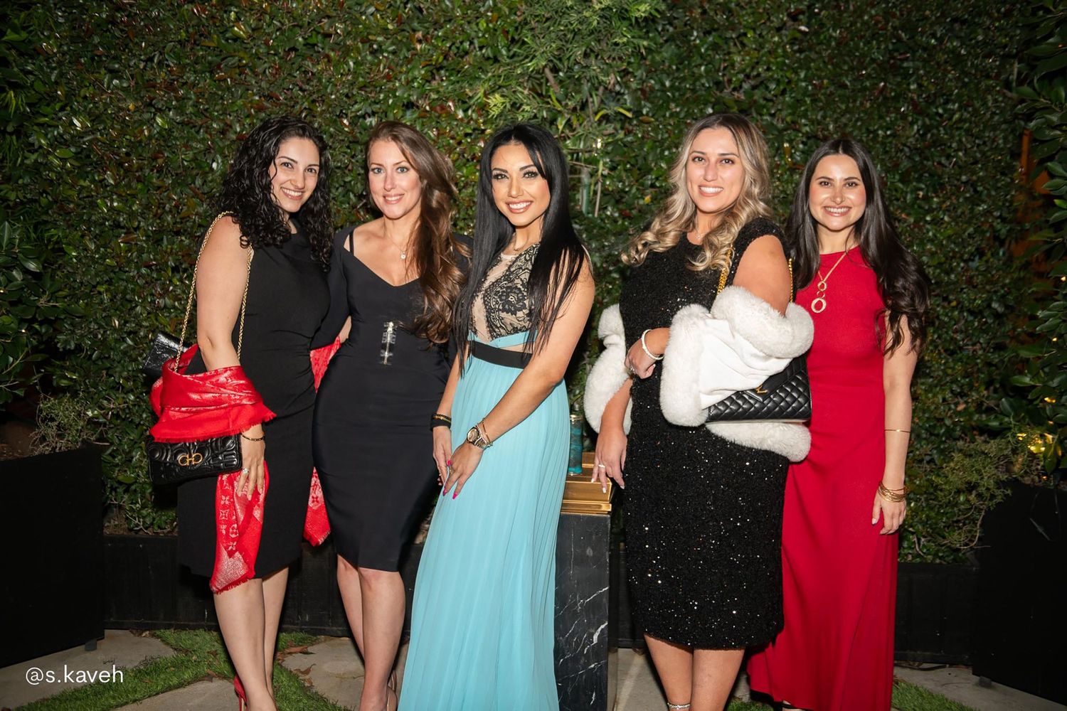 Five women in elegant evening dresses in black, red and turquoise pose together at a formal event in front of a lush green hedge wall.