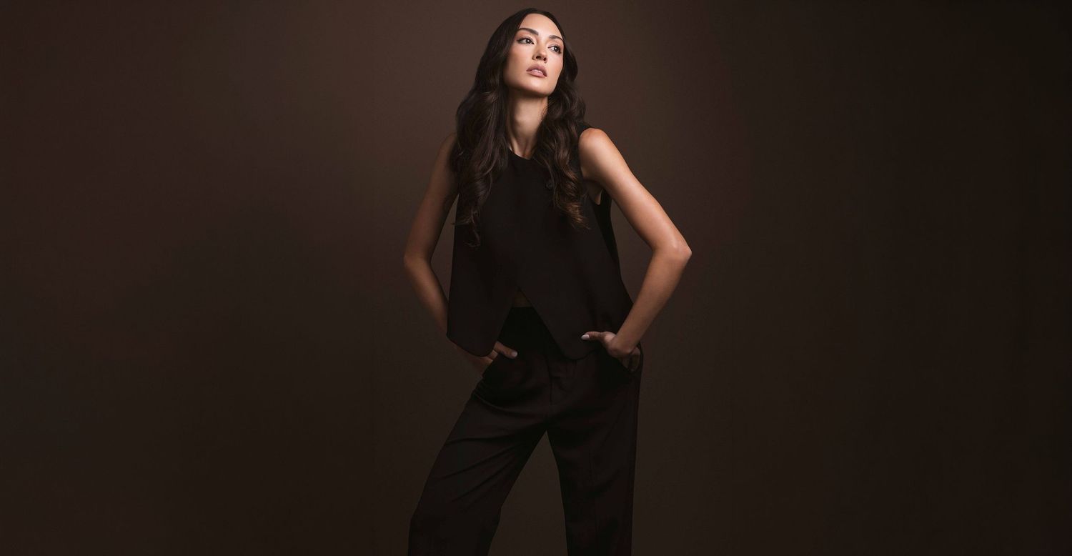 Stylish woman in black sleeveless blouse and wide-leg pants poses against dark brown background.