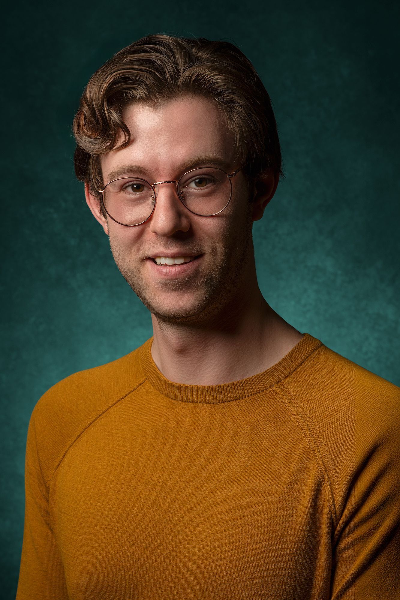 Young smiling man with round glasses and curly hair wearing mustard sweater poses for portrait.