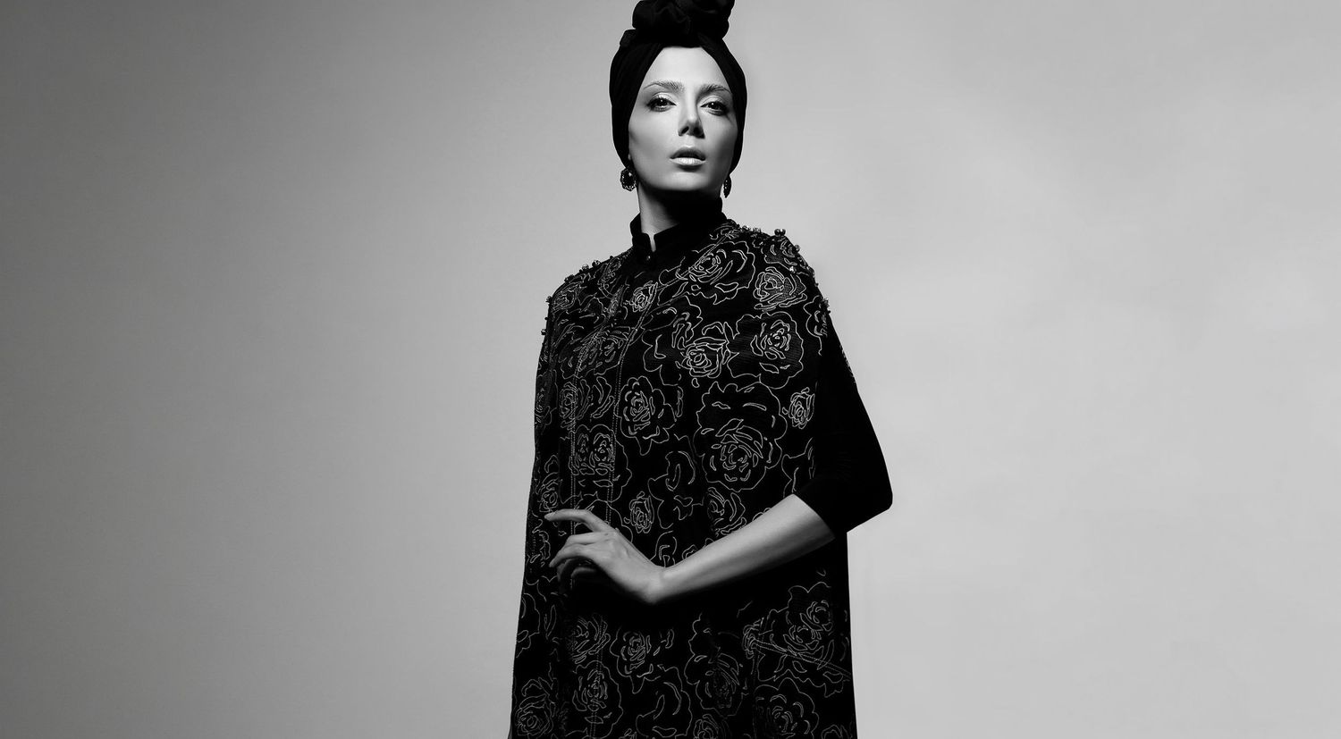 Woman in black embroidered dress with dramatic updo poses against gray background in monochrome fashion portrait.