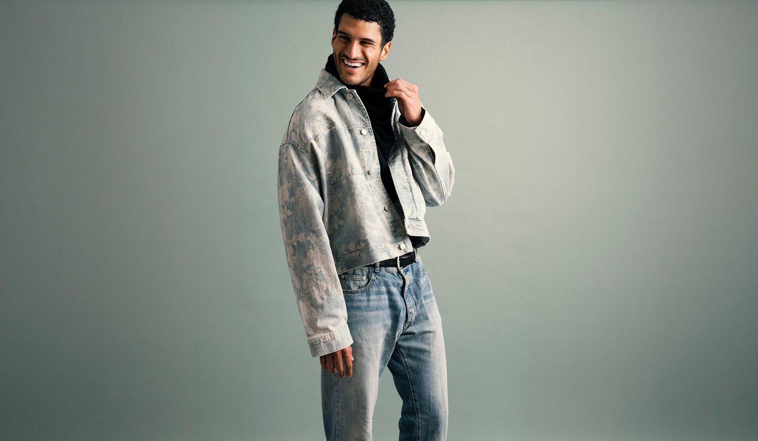 Young man in light gray cropped jacket and wide-leg jeans smiles in casual fashion studio shoot.