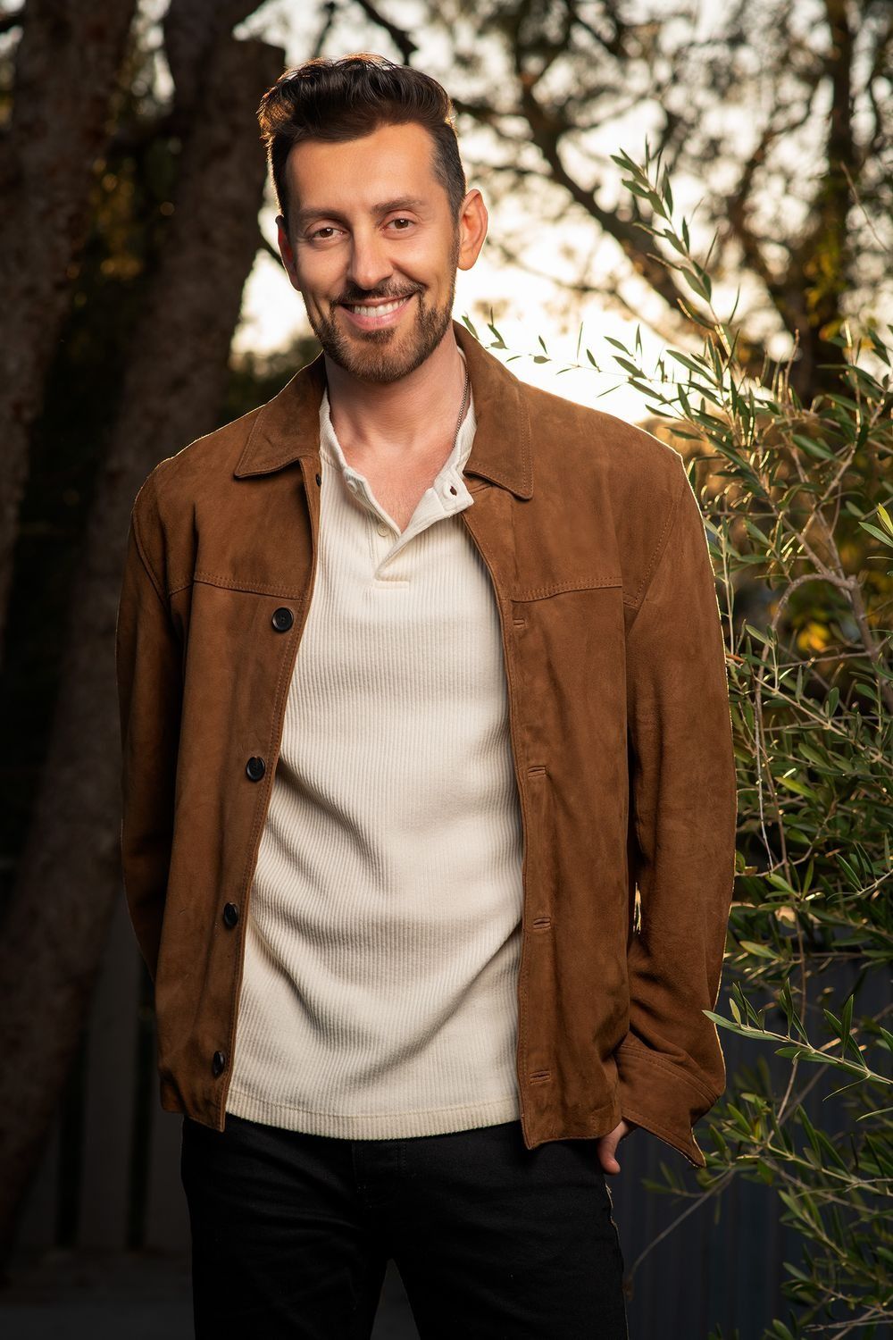 Smiling bearded man in brown suede jacket and cream sweater poses outdoors near trees.