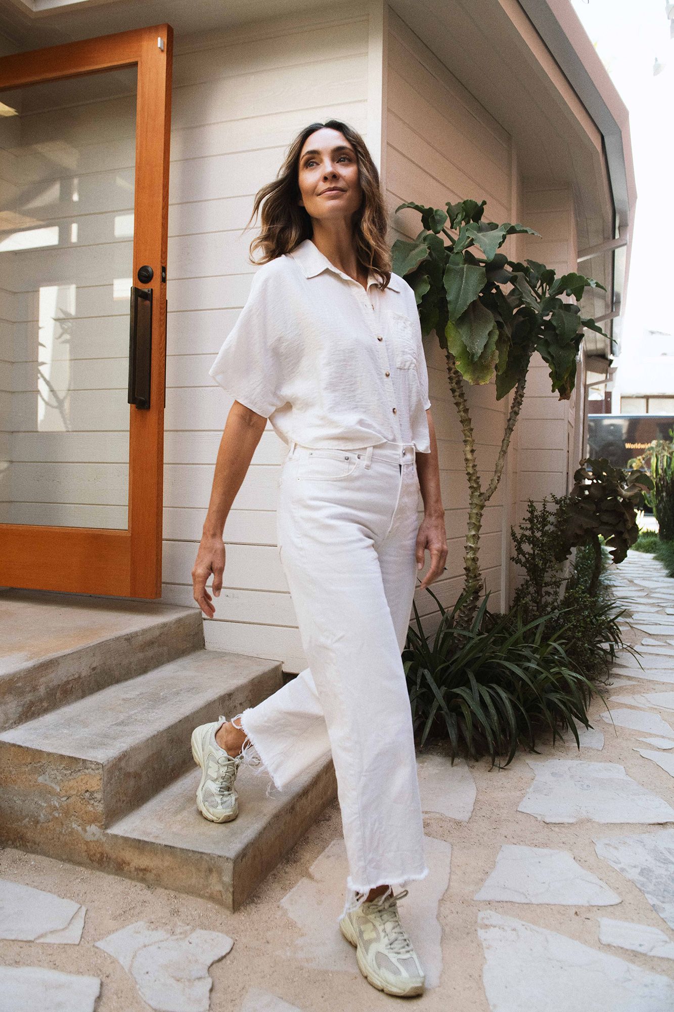 Woman in white linen shirt and wide-leg jeans with chunky sneakers standing outside a modern home.