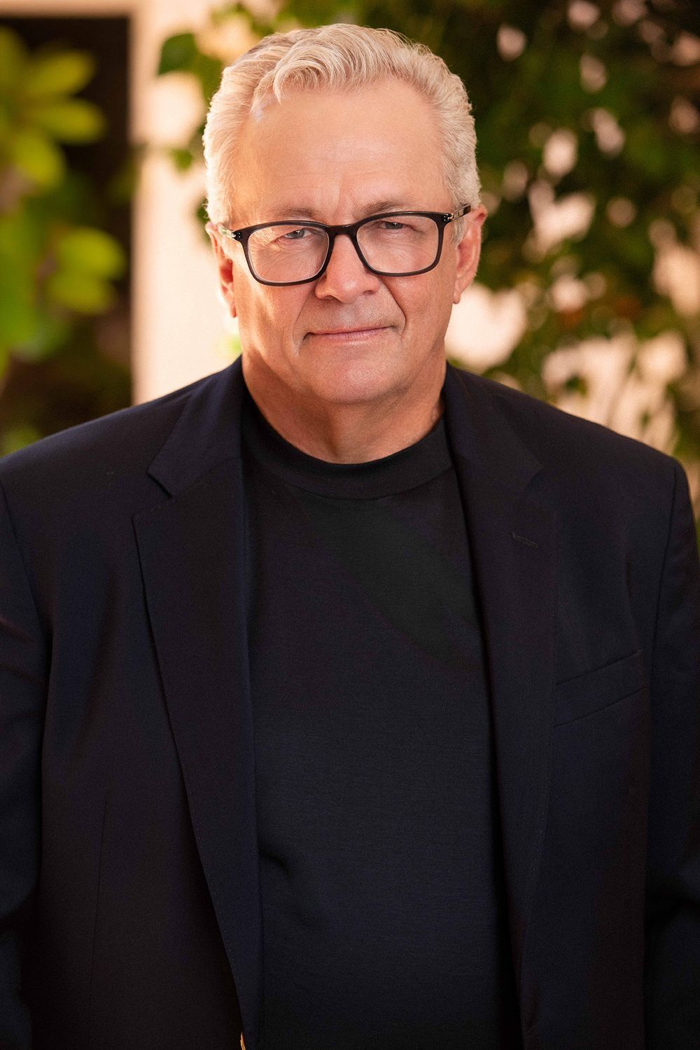 Senior professional man with white hair and glasses in black blazer, outdoor headshot.