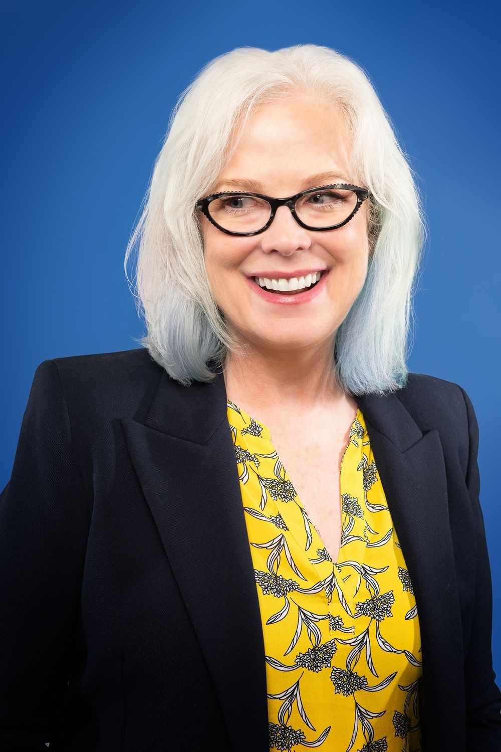 Smiling older woman with white hair and glasses in a yellow blouse and black blazer.