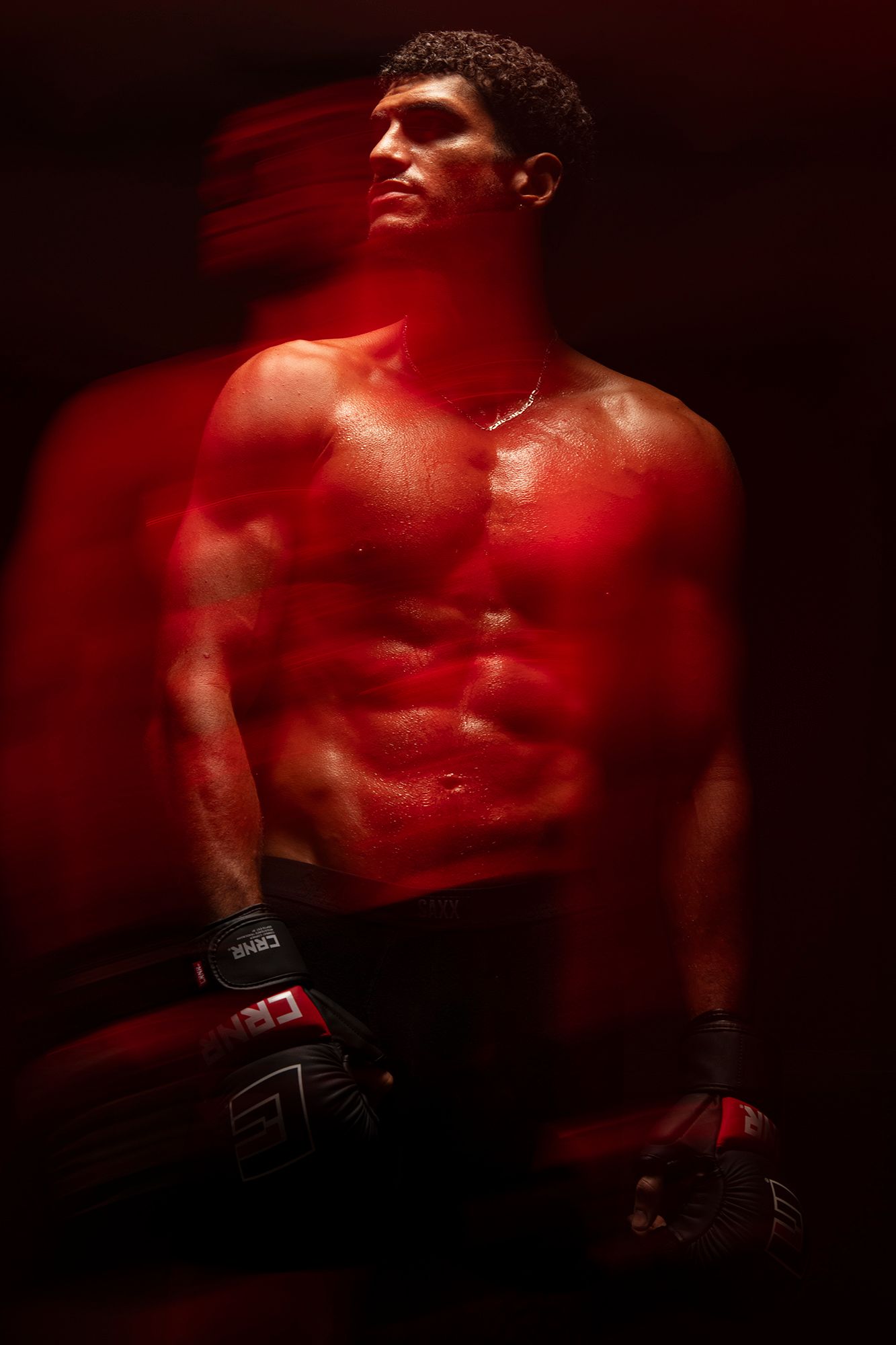 Dramatic red-lit photo of a muscular shirtless male fighter wearing boxing gloves in dark studio setting.