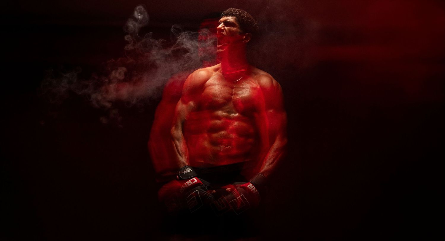 Muscular shirtless man posed dramatically in red-lit smoke holding a drink in dark studio setting.