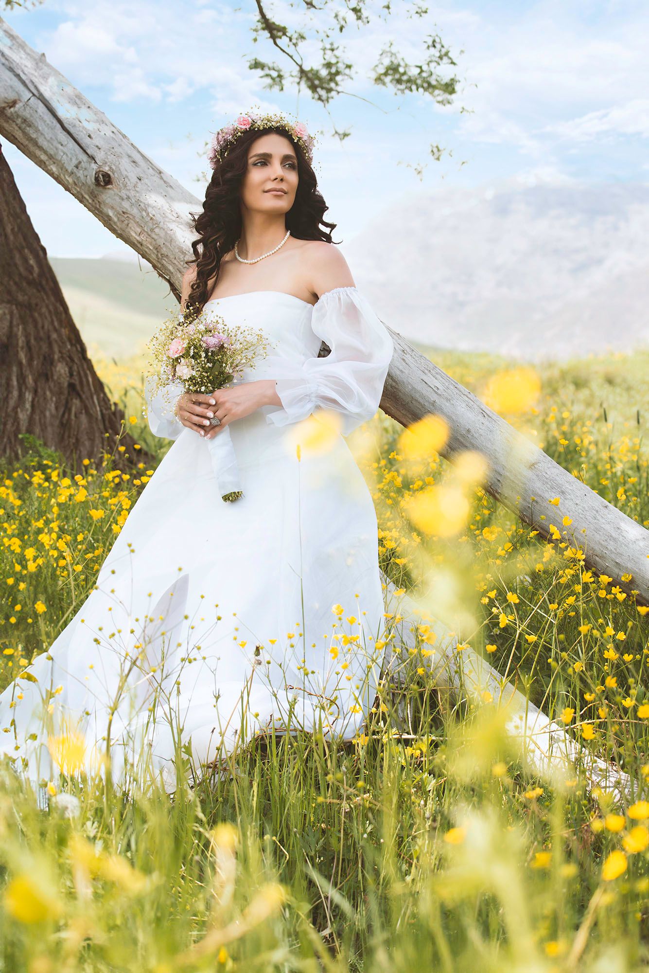 Bride in white off-shoulder gown holding wildflower bouquet standing in yellow flower field with mountains backdrop.