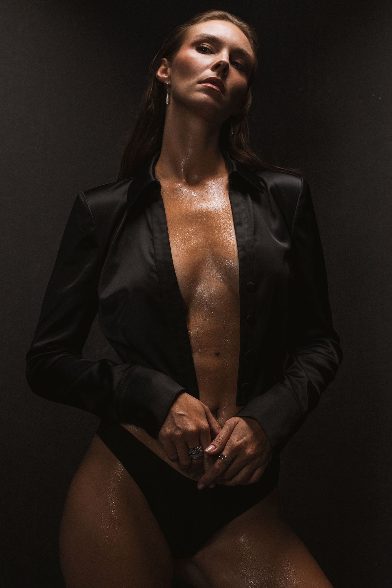 Woman in wet black blazer posing dramatically against dark background in moody fashion photography.