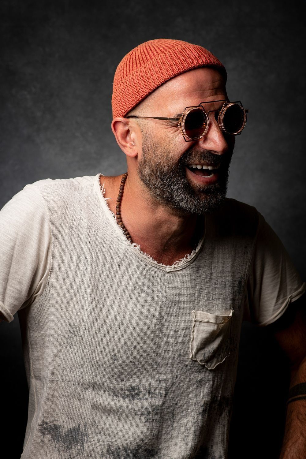 Bearded man in orange beanie and round sunglasses laughing in studio.