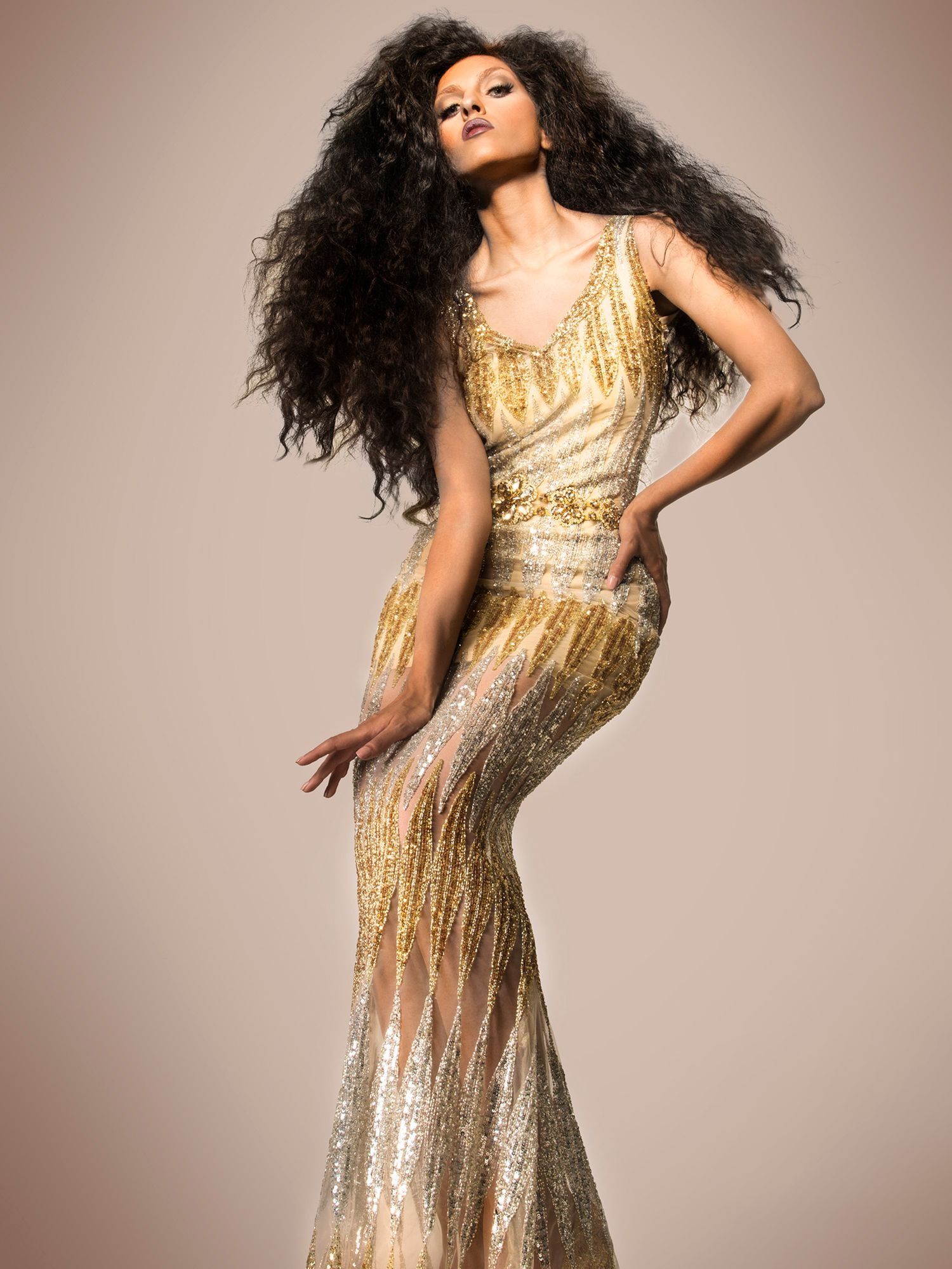 A model in a gold sequined mermaid gown poses with voluminous curly black hair in a fashion shoot.