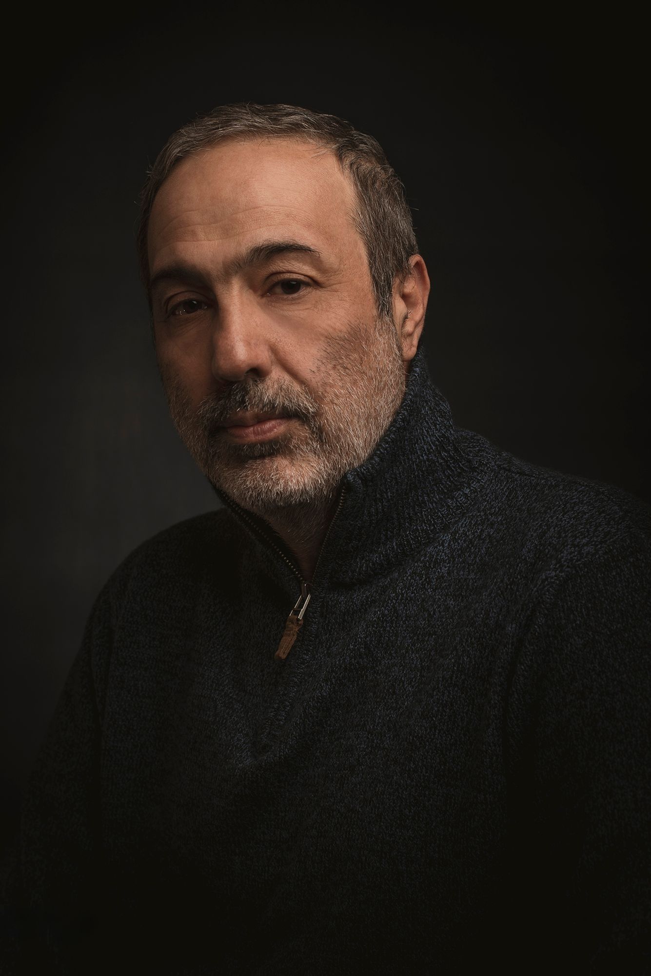 Middle-aged man with salt-and-pepper beard wearing dark zip-up sweater poses against moody dark background.