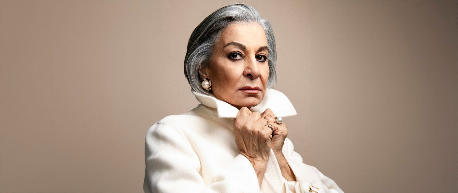 Elegant older woman with silver hair in white turtleneck, posing confidently against beige background.