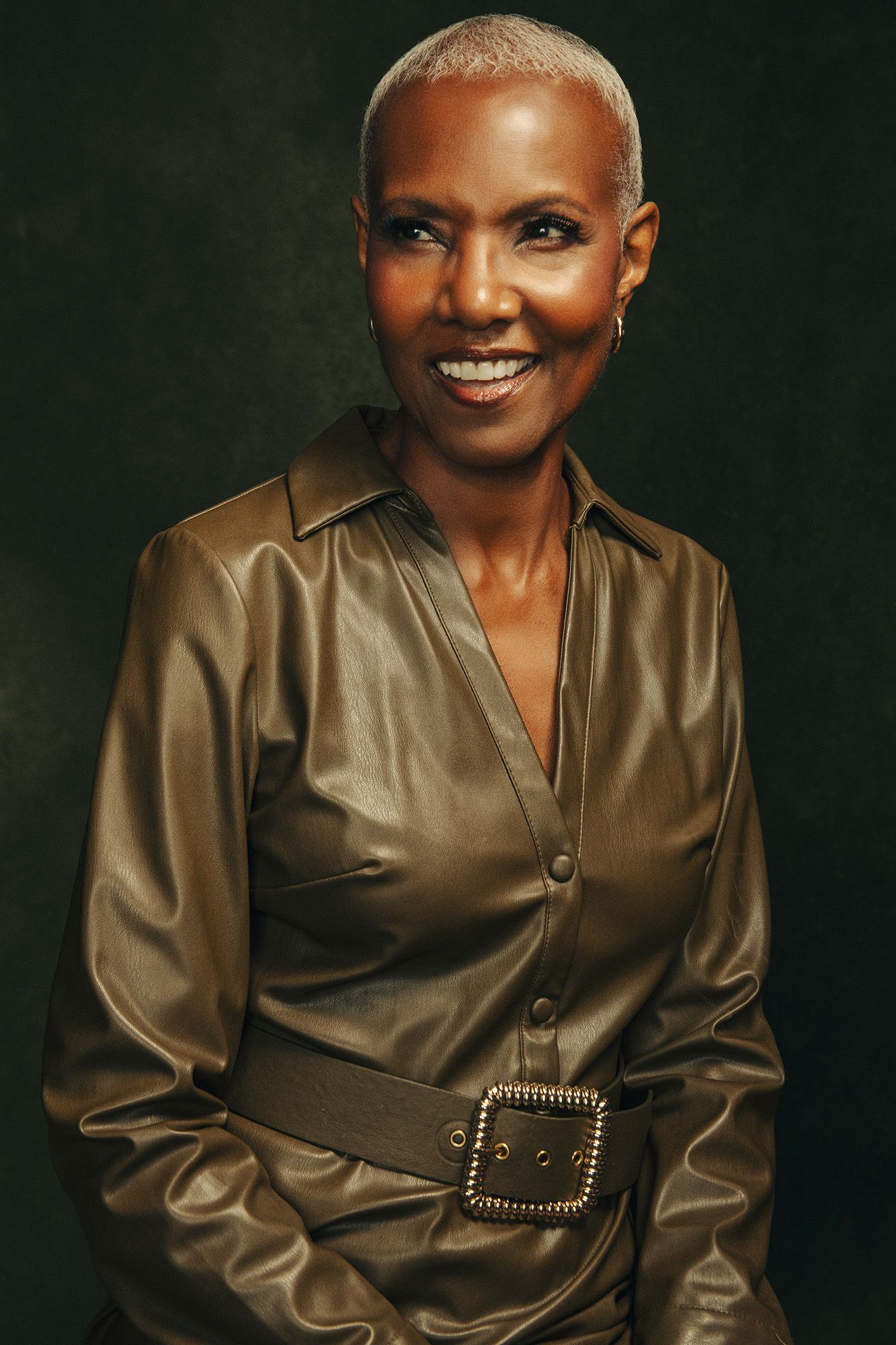 Smiling woman with short blonde hair wearing a gold leather belted shirt dress poses against a dark green background.