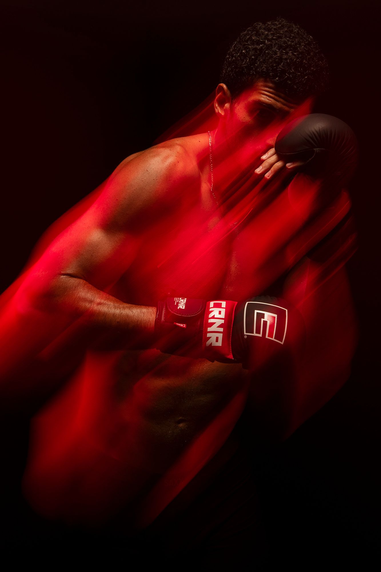 Muscular boxer in red dramatic lighting wearing RNR boxing gloves in a powerful defensive stance against dark background.