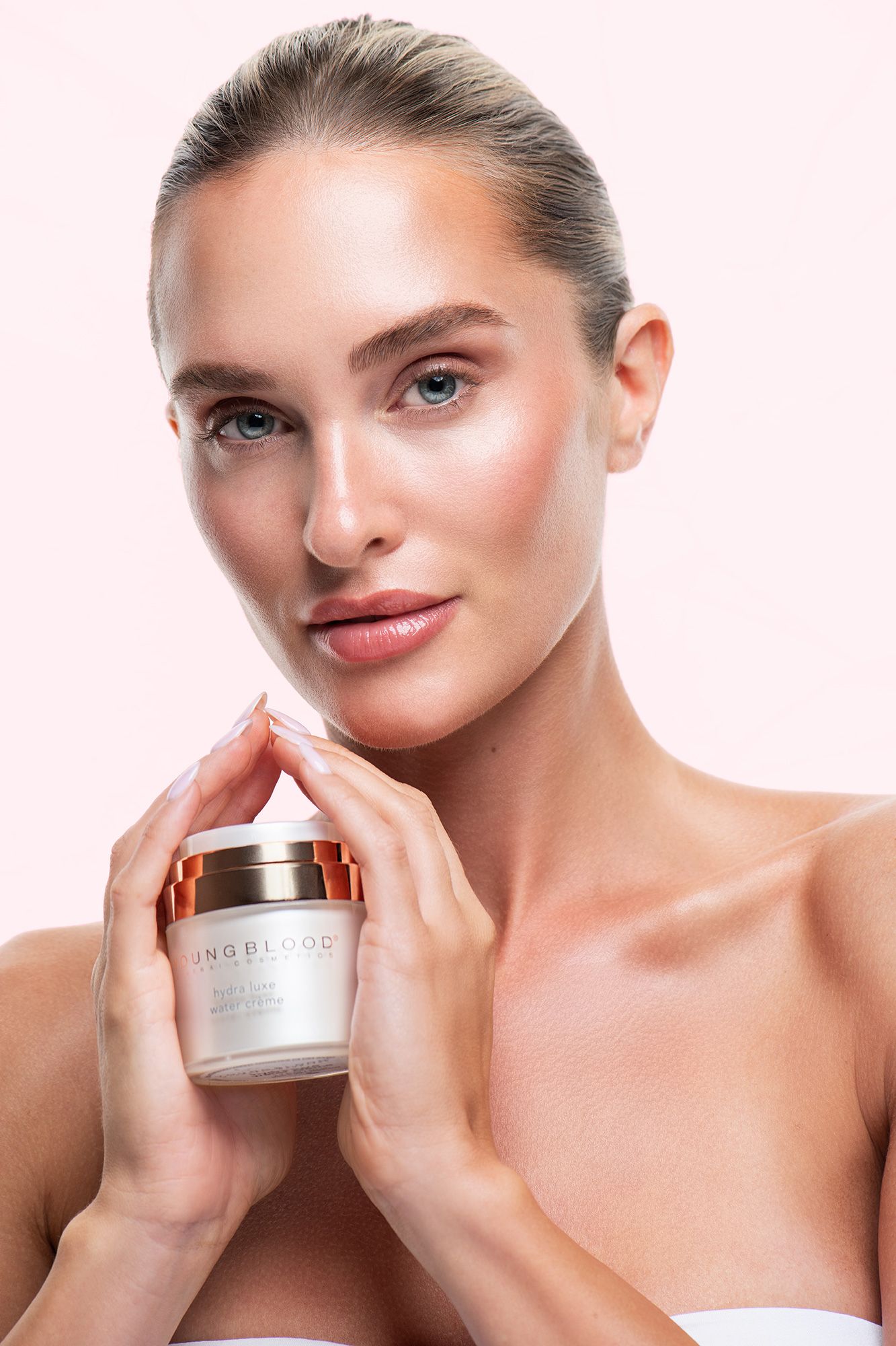 Woman with glowing skin holding a Youngblood skincare cream jar, showcasing a radiant complexion against a soft pink background.