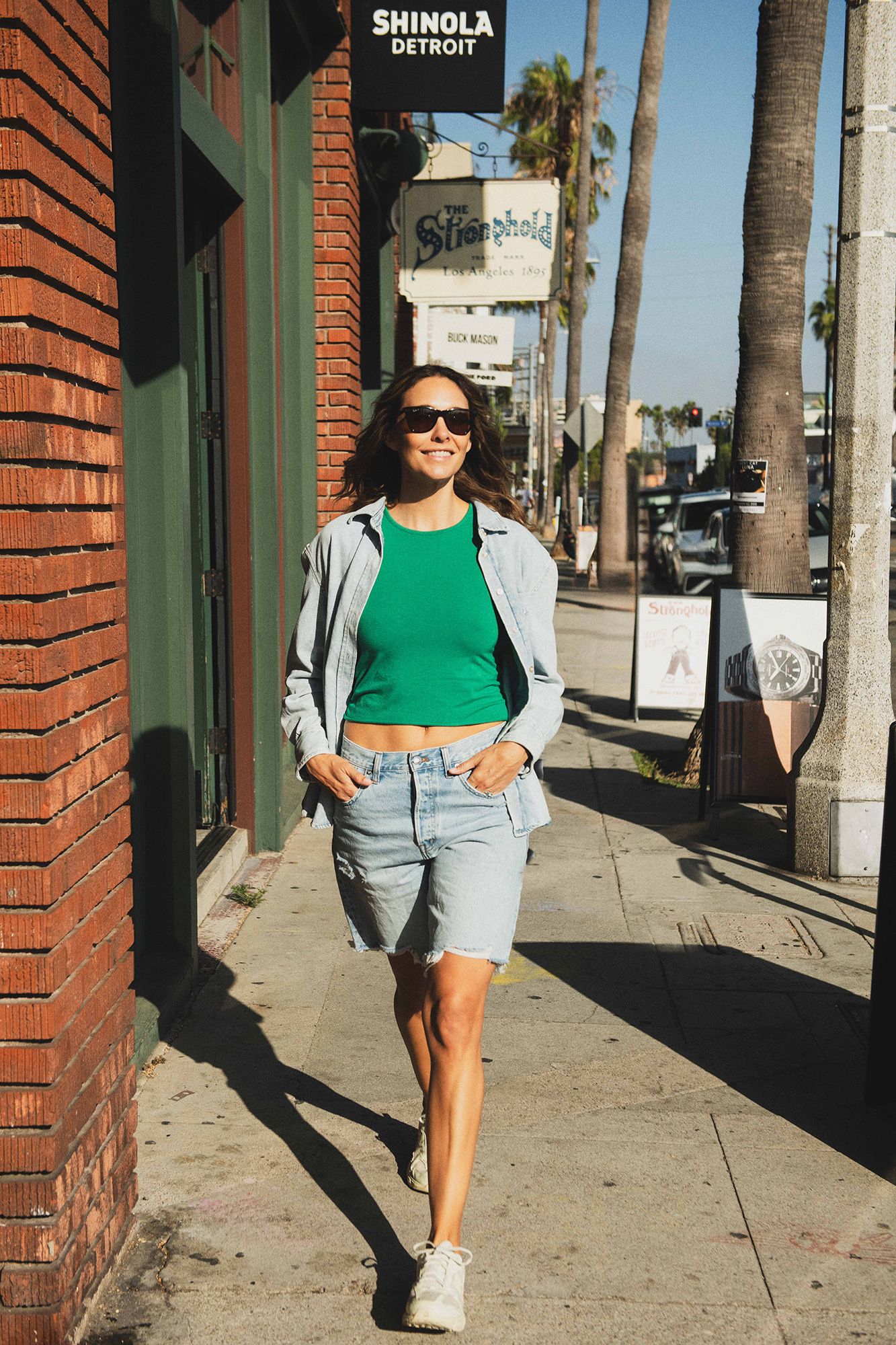 Woman in green crop top, denim shorts, and gray hoodie walking on sunny sidewalk near Shinola Detroit store.
