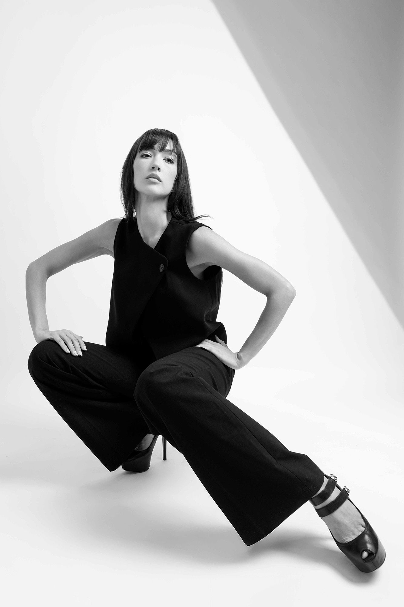 Black and white fashion photo of model in wide-leg pants seated against minimalist studio backdrop.