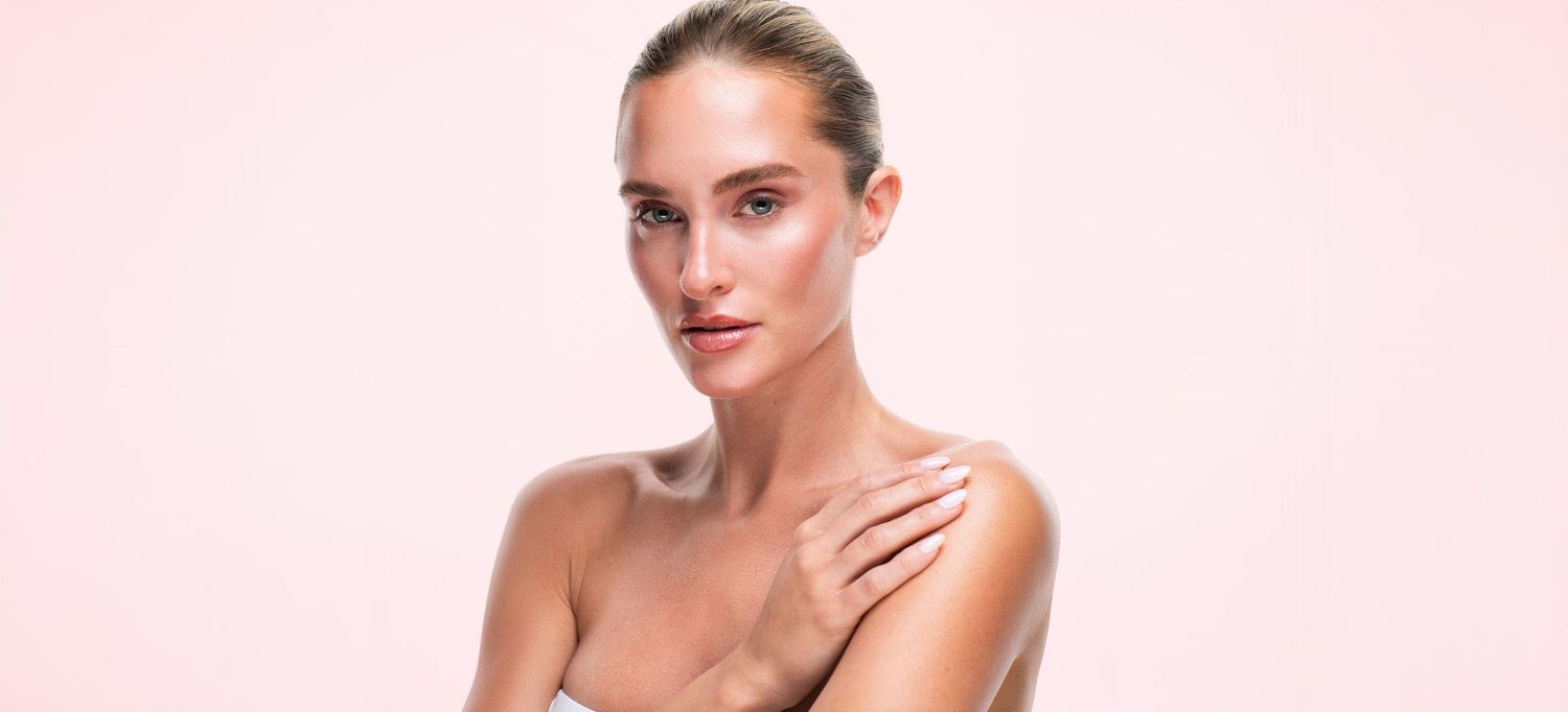 Woman with glowing, radiant skin and slicked-back hair posing with one hand on her bare shoulder against a soft pink background.