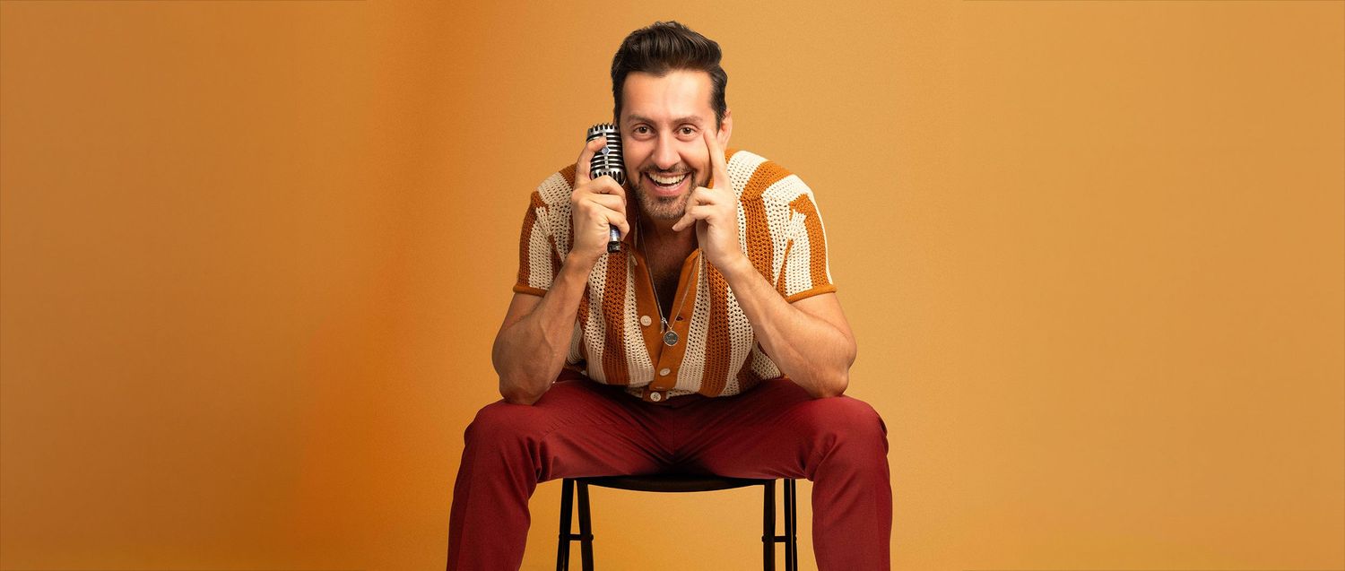 Smiling man in retro striped shirt sitting on stool holding phone against orange background.