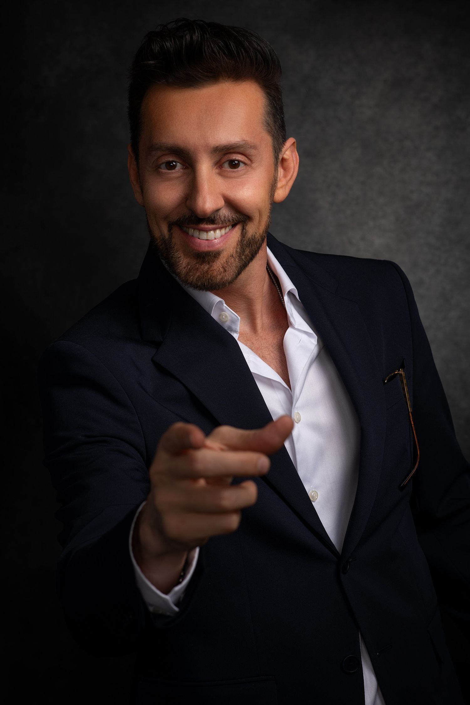 Cheerful bearded man in navy suit pointing at camera smiles during professional studio photo session.